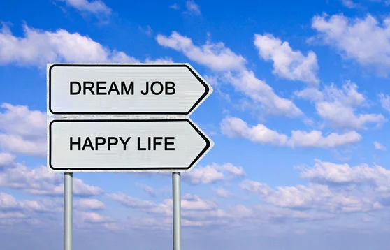 A signpost with two directional arrows pointing opposite ways, labeled “Dream Job” and “Happy Life,” against a bright blue sky with scattered clouds.